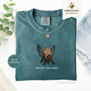 Peut inclure: T-shirt bleu épicéa avec un motif brodé d'un paresseux chevauchant un papillon de nuit, avec le texte "PROTECT OUR PARKS". Le t-shirt a un col rond et des manches courtes. Un collier doré avec un pendentif est également visible.