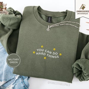 May include: An olive green sweatshirt with the embroidered phrase "YOU CAN DO HARD THINGS" in white and yellow stars. A silver necklace is draped across the front. The sweatshirt is folded on top of blue denim jeans.