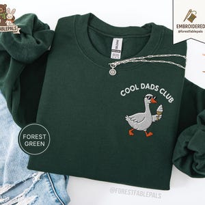 May include: Forest green sweatshirt with the text "COOL DADS CLUB" and an embroidered goose wearing sunglasses and holding an ice cream cone. A silver necklace is draped across the sweatshirt. The sweatshirt is folded on top of a pair of blue jeans.