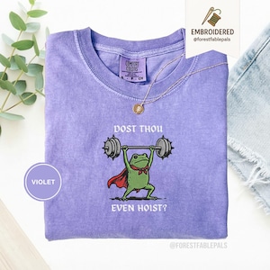 May include: A violet Comfort Colors t-shirt with an embroidered frog lifting weights. The frog wears a red cape and a green bow tie. The text on the shirt reads "DOST THOU EVEN HOIST?" A gold necklace and a small tag are also visible.