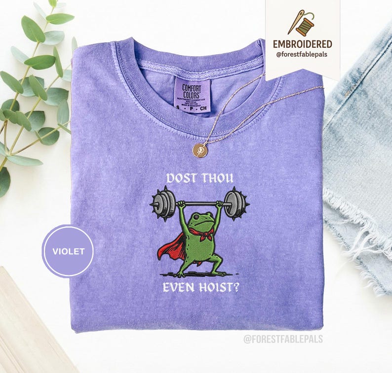 May include: A violet Comfort Colors t-shirt with an embroidered frog lifting weights. The frog wears a red cape and a green bow tie. The text on the shirt reads "DOST THOU EVEN HOIST?" A gold necklace and a small tag are also visible.