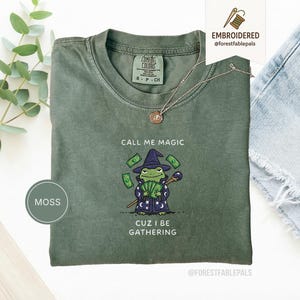 Puede incluir: Camiseta verde musgo con un diseño de rana mago. La rana lleva un sombrero y una túnica de mago, sosteniendo dinero y un bastón. El texto en la camiseta dice "CALL ME MAGIC CUZ I BE GATHERING". Marca Comfort Colors, talla S-P-CH.