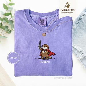May include: A violet Comfort Colors t-shirt with an embroidered otter design. The otter is wearing a red cape and holding a sword, with the word "Huzzah" below. A gold necklace and a "Violet" circle are also visible.