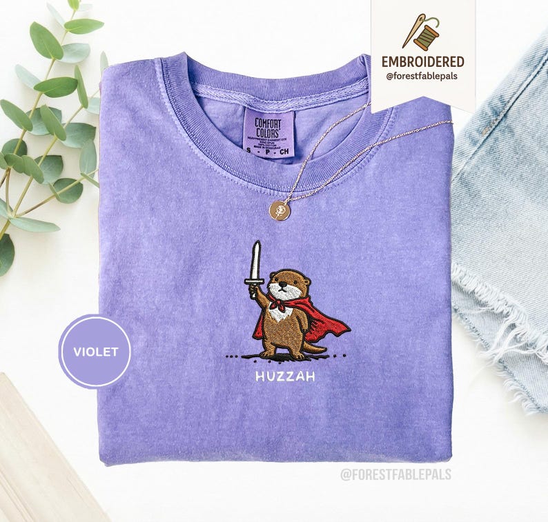 May include: A violet Comfort Colors t-shirt with an embroidered otter design. The otter is wearing a red cape and holding a sword, with the word "Huzzah" below. A gold necklace and a "Violet" circle are also visible.