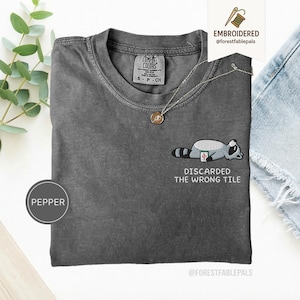 Pode incluir: Camiseta cinza escuro com gola redonda e mangas curtas. Apresenta um design bordado de guaxinim e o texto "DISCARDED THE WRONG TILE". Um colar de prata com um pingente também é visível.