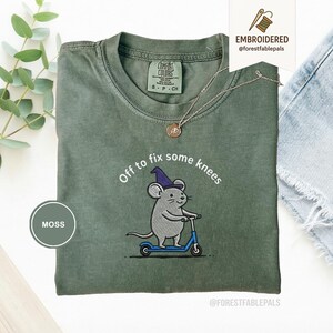 May include: Moss green t-shirt with an embroidered design of a gray mouse wearing a purple wizard hat and riding a blue scooter. The text "Off to fix some knees" is above the mouse. The shirt has a crew neck and short sleeves.