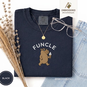 May include: A navy blue t-shirt with the word "FUNCLE" embroidered in white above a brown bear wearing sunglasses and holding an ice cream cone. A gold necklace and clear glasses are on the shirt. The shirt is labeled "BLACK".
