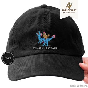 Puede incluir: Una gorra de béisbol de pana negra con un bordado que muestra un gato marrón con gafas de sol montado sobre un dinosaurio azul con detalles rosas. Debajo del gráfico, el texto dice THIS IS AN OUTRAGE en letras mayúsculas blancas.