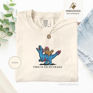 May include: Ivory-colored t-shirt with an embroidered design of a brown cat wearing sunglasses riding a blue dinosaur. The text "THIS IS AN OUTRAGE" is below the design. The shirt has a crew neck and short sleeves.