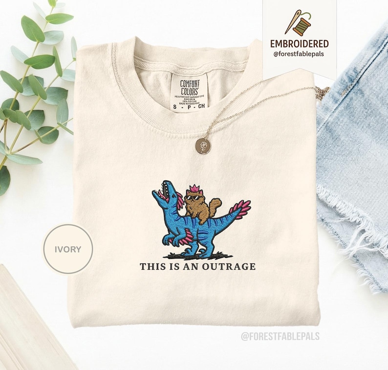 May include: Ivory-colored t-shirt with an embroidered design of a brown cat wearing sunglasses riding a blue dinosaur. The text "THIS IS AN OUTRAGE" is below the design. The shirt has a crew neck and short sleeves.