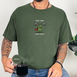 May include: A moss green t-shirt with a cartoon frog lifting weights. The frog is depicted with a red cape and bow tie, and the text "DOST THOU EVEN HOIST?" is printed above and below. The shirt is a solid colour.