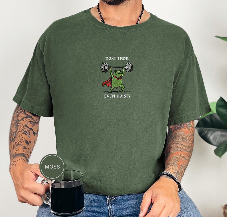 May include: A moss green t-shirt with a cartoon frog lifting weights. The frog is depicted with a red cape and bow tie, and the text "DOST THOU EVEN HOIST?" is printed above and below. The shirt is a solid colour.