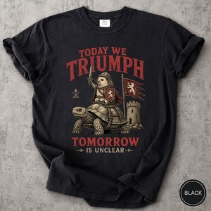 Może przedstawiać: Czarny t-shirt z krótkim rękawem z grafiką jeża w zbroi jadącego na żółwiu. Napis "TODAY WE TRIUMPH" znajduje się powyżej, a "TOMORROW IS UNCLEAR" poniżej. Wzór zawiera kamienną wieżę, flagę z lwem oraz skrzyżowane miecze.
