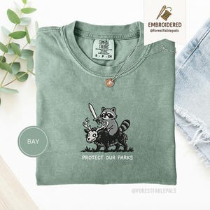 Może przedstawiać: Szałwiowa zielona koszulka Comfort Colors z haftowanym wzorem. Wzór przedstawia szopa trzymającego miecz, jadącego na ciemnym zwierzęciu z rogami, z napisem "PROTECT OUR PARKS" poniżej. Srebrny naszyjnik z wisiorkiem znajduje się na koszulce.