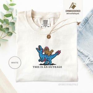 May include: White Comfort Colors t-shirt with an embroidered design. The design features a brown cat wearing sunglasses riding a blue dinosaur with pink accents. The text "THIS IS AN OUTRAGE" is below the design.