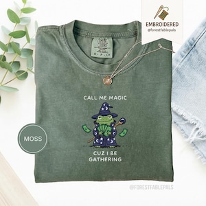 May include: Moss green t-shirt with the text "CALL ME MAGIC CUZ I BE GATHERING". The shirt features an embroidered cartoon frog wearing a wizard hat and robe, holding a wand and money. The shirt has a crew neck and short sleeves.