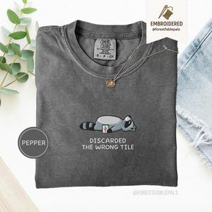 Peut inclure: Un t-shirt gris avec un col rond et des manches courtes. Le t-shirt présente un motif de raton laveur brodé avec le texte "DISCARDED THE WRONG TILE". Un collier en or avec un pendentif est sur le t-shirt.