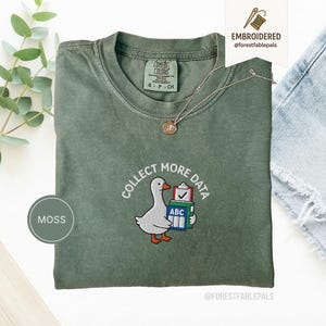 Op de afbeelding: Mosgroen Comfort Colors T-shirt met een geborduurd ontwerp. Het ontwerp toont een cartoon gans die boeken draagt en de tekst "COLLECT MORE DATA". Een zilveren ketting met een hanger is over het T-shirt gedrapeerd.