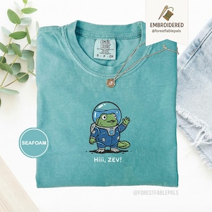 May include: Seafoam green t-shirt featuring an embroidered cartoon frog in a spacesuit. The frog is waving, with the text "Hiii, ZEV!" beneath. The shirt has a crew neck and the brand "Comfort Colors" is visible.