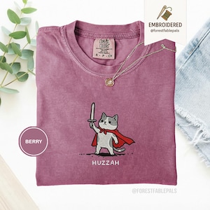Puede incluir: Camiseta color bayas con un diseño de gato bordado. El gato, con una capa roja y una espada, está sobre una línea de puntos. La palabra "Huzzah" está debajo del gato. Un collar dorado está sobre la camiseta.