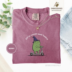 Puede incluir: Camiseta color bayas con cuello redondo. La camiseta presenta un bordado de una rana verde con un sombrero de mago montada en un scooter, con el texto "Off to commit aunt foolery". Un collar dorado está sobre la camiseta.