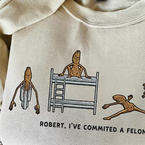 Puede incluir: Sudadera de color crema con un diseño bordado que presenta vegetales de raíz antropomórficos en varias poses. Uno está en una litera, otro en un tubo y otros en poses de acción. El texto "ROBERT, I'VE COMMITTED A FELONY" está debajo.