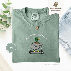 Op de afbeelding: Saliegroen T-shirt met de geborduurde tekst "Off to commit dad foolery" en een cartoon eend op een scooter. Het shirt heeft een ronde hals en korte mouwen. Het merklabel "Comfort Colors" is zichtbaar.
