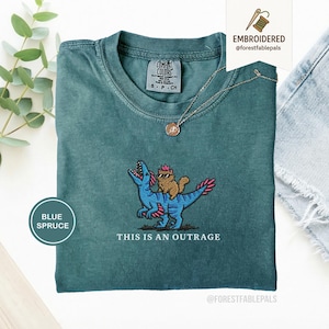 May include: Blue spruce t-shirt with an embroidered design of a cat riding a blue dinosaur. The text "THIS IS AN OUTRAGE" is below the design. The shirt has a crew neck and short sleeves.