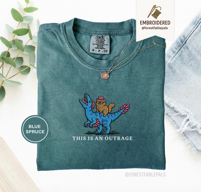May include: Blue spruce t-shirt with an embroidered design of a cat riding a blue dinosaur. The text "THIS IS AN OUTRAGE" is below the design. The shirt has a crew neck and short sleeves.