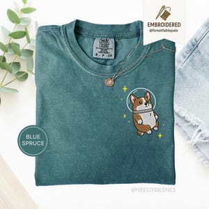 Könnte beinhalten: Blau-Fichte-T-Shirt mit einem gestickten Cartoon-Corgi, der einen Weltraumhelm trägt. Das T-Shirt hat einen Rundhalsausschnitt und kurze Ärmel. Der Corgi ist braun und weiß mit einem weißen Weltraumhelm und gelben Sternen. Das T-Shirt hat den Text "COMFORT COLORS".