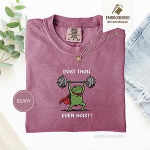 May include: Berry-coloured t-shirt with a crew neck. The shirt features a graphic of a green frog lifting weights, wearing a red cape and bow tie. The text "DOST THOU EVEN HOIST?" is printed below the frog. A gold necklace is draped over the shirt.
