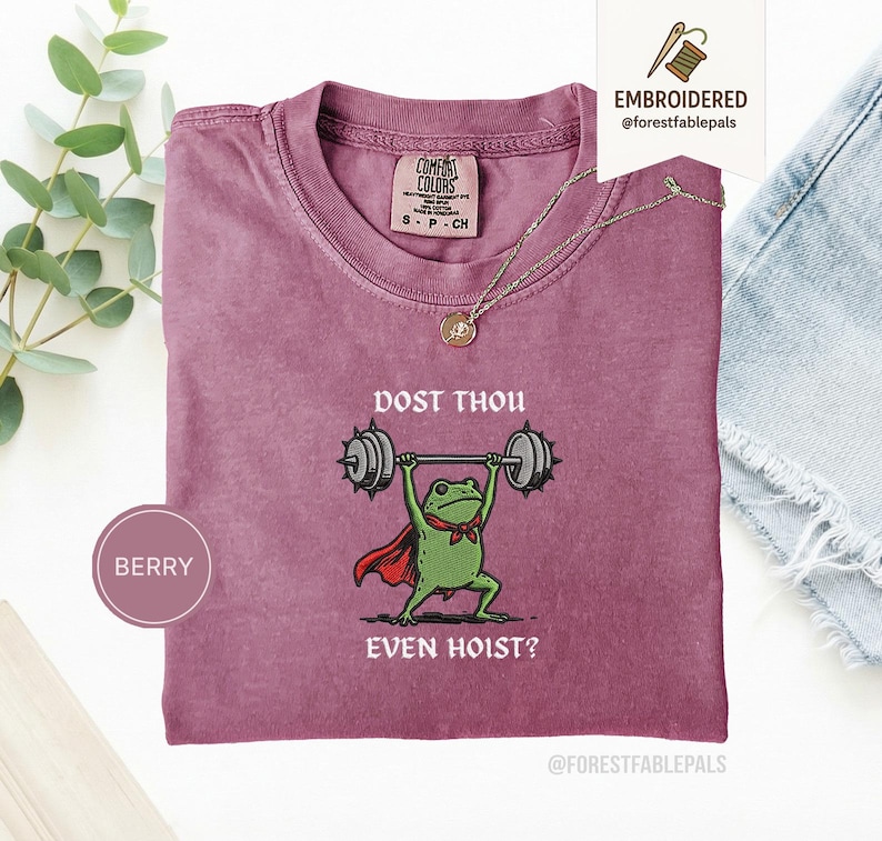 May include: Berry-coloured t-shirt with a crew neck. The shirt features a graphic of a green frog lifting weights, wearing a red cape and bow tie. The text "DOST THOU EVEN HOIST?" is printed below the frog. A gold necklace is draped over the shirt.