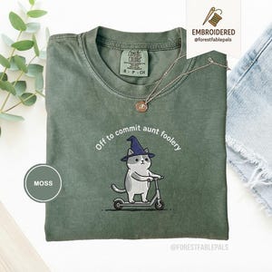 Puede incluir: Camiseta verde musgo con la frase bordada "Off to commit aunt foolery" y un gato en un patinete, con un sombrero de bruja. La camiseta tiene cuello redondo y mangas cortas. También es visible un collar plateado con un colgante.