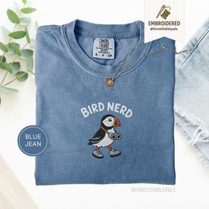 Op de afbeelding: Een blauw spijkerkleurig t-shirt met een geborduurde afbeelding van een papegaaiduiker met een verrekijker en grijze schoenen, met de tekst BIRD NERD er in een boog boven. Het Comfort Colors label geeft maat S aan en is 100% katoen.