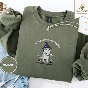 Puede incluir: Una sudadera verde oliva con la frase bordada "Off to commit aunt foolery". El diseño presenta un gato con un sombrero de bruja montado en un scooter. Un collar plateado está drapeado sobre la sudadera.