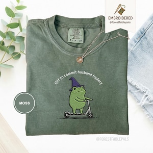 Op de afbeelding: Mosgroen T-shirt met een geborduurde kikker met een heksenhoed op een scooter. De tekst "Off to commit husband foolery" staat erboven. Een zilveren ketting met hanger is zichtbaar.