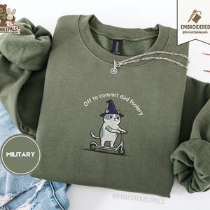 Könnte beinhalten: Olivgrünes Sweatshirt mit dem gestickten Schriftzug "Off to commit dad foolery" und einer Katze mit Hexenhut auf einem Roller. Eine silberne Halskette mit Sonnenanhänger ist zu sehen.