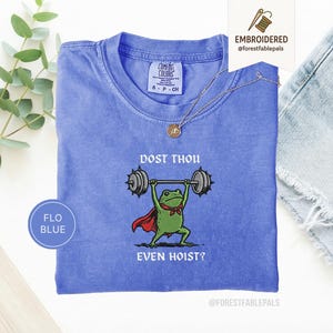 May include: A blue Comfort Colors t-shirt featuring a frog lifting weights. The frog is depicted with a red cape and a green bow tie. The text on the shirt reads "DOST THOU EVEN HOIST?" and "FLO BLUE".
