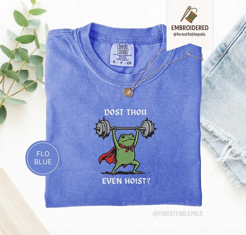 May include: A blue Comfort Colors t-shirt featuring a frog lifting weights. The frog is depicted with a red cape and a green bow tie. The text on the shirt reads "DOST THOU EVEN HOIST?" and "FLO BLUE".