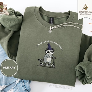 May include: An olive green sweatshirt with the embroidered phrase "Off to commit husband foolery" and a raccoon wearing a wizard hat riding a scooter. A silver necklace with a sun pendant is draped across the sweatshirt.