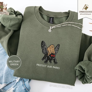 Peut inclure: Sweat-shirt vert militaire avec un motif brodé d'une créature brune chevauchant un papillon noir, avec le texte "PROTECT OUR PARKS". Le sweat-shirt a un col rond et des manches longues. Un collier argenté avec un pendentif est sur le sweat-shirt.