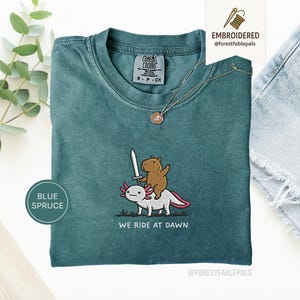 Peut inclure: T-shirt bleu épicéa avec un motif brodé d'un capybara maniant une épée sur un axolotl. Le texte "WE RIDE AT DAWN" est en dessous. Le t-shirt a un col rond et des manches courtes.