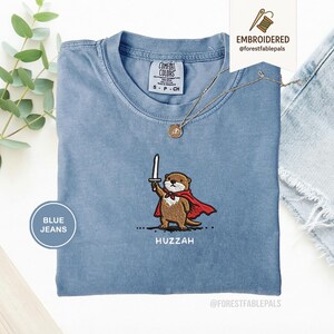 May include: A light blue t-shirt with an embroidered otter wearing a red cape and holding a sword. The text "Huzzah" is below the otter. The shirt has a small Comfort Colors tag and a gold necklace. The words "Embroidered" and "Blue Jeans" are also visible.