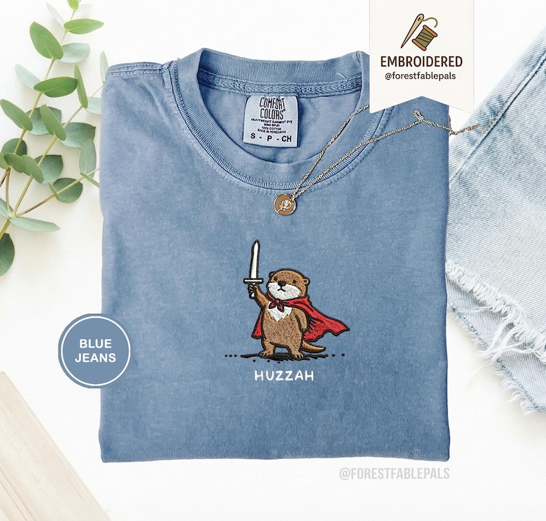 May include: A light blue t-shirt with an embroidered otter wearing a red cape and holding a sword. The text "Huzzah" is below the otter. The shirt has a small Comfort Colors tag and a gold necklace. The words "Embroidered" and "Blue Jeans" are also visible.