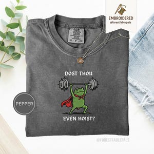 Może przedstawiać: Szara koszulka Comfort Colors z grafiką żaby podnoszącej ciężary. Żaba ma czerwoną pelerynę i muszkę, z napisem "DOST THOU EVEN HOIST?" nad i pod żabą. Złoty naszyjnik jest na koszulce.