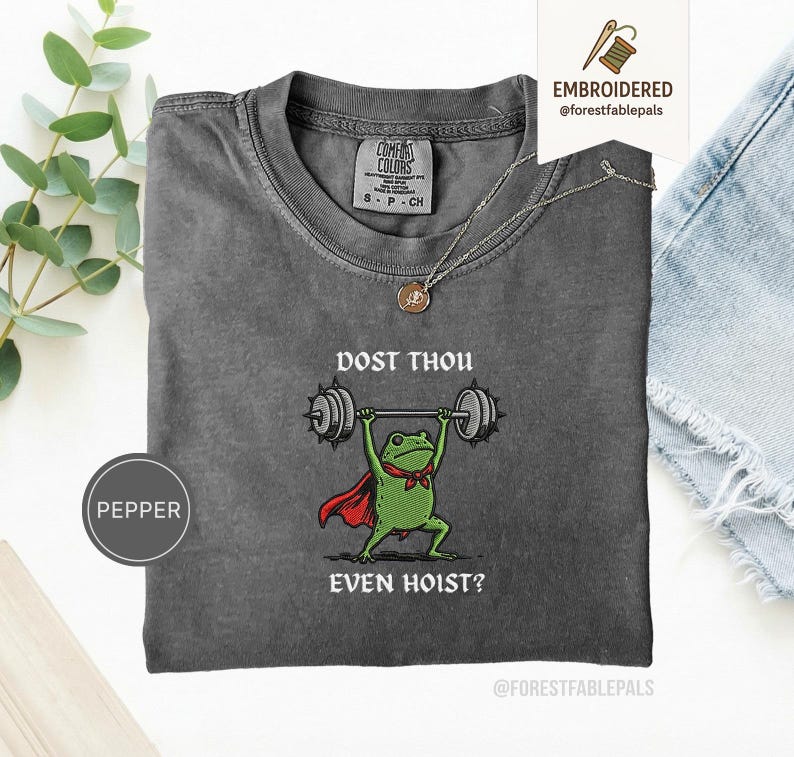May include: A grey Comfort Colors t-shirt featuring a frog lifting weights. The frog is depicted with a red cape and bow tie, with the text "DOST THOU EVEN HOIST?" above and below. A gold necklace is draped on the shirt.