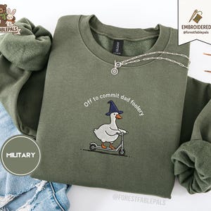 Op de afbeelding: Een olijfgroene sweatshirt met de geborduurde tekst "Off to commit dad foolery" en een gans met een tovenaars hoed op een scooter. Een zilveren ketting is over de sweatshirt gedrapeerd.