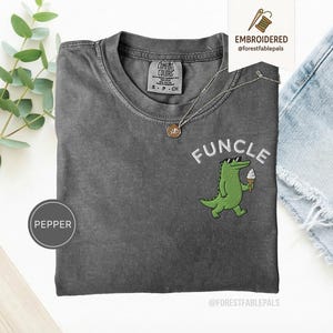 May include: A grey Comfort Colors t-shirt featuring the embroidered word "FUNCLE" in white, and a green alligator wearing sunglasses and holding an ice cream cone. A silver necklace with a pendant is on the shirt.