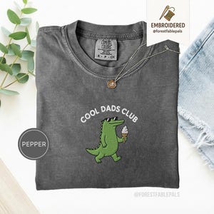 May include: A gray Comfort Colors t-shirt with the embroidered phrase "COOL DADS CLUB" and a green alligator wearing sunglasses and holding an ice cream cone. A gold necklace and a small tag are also visible.