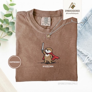 May include: Espresso-colored t-shirt with an embroidered otter design. The otter, wearing a red cape and holding a sword, is embroidered above the word "Huzzah". The shirt has a crew neck and short sleeves. A silver necklace with a pendant is also visible.